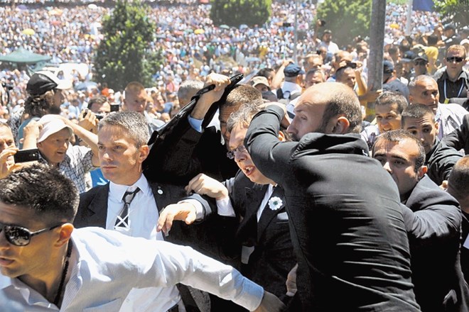 Varnostniki ščitijo srbskega premierja Vučića pred letečimi predmeti in jeznimi ljudmi v množici. 