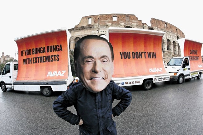 Igralec z masko Silvia Berlusconija stoji pred tovornjakom s tremi plakati, ki Berlusconiju sporočajo: Če se spečaš s...