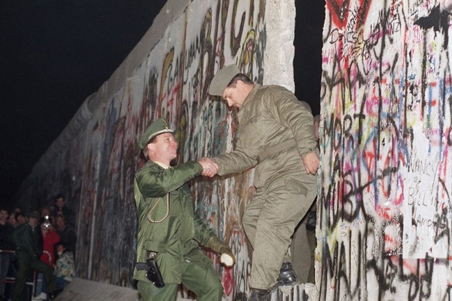 Zahodnonemški policist z dlanjo pomaga vzhodnonemškemu varuhu meje vstopiti skozi luknjo v Berlinskem zidu.