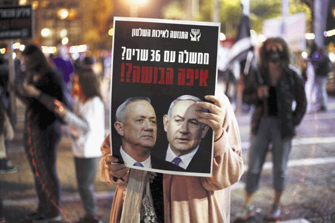 Izraelka  nosi plakat s fotografijama   Netanjahuja in Ganca ter se sprašuje, ali vlada s kar 36 ministri ni sramota za...