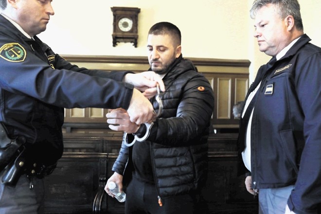 Mehmeda Mahmutovića na sodišče spremljajo  trije pravosodni policisti. Ima vklenjene roke in noge, lisice na rokah mu med...
