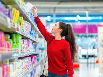 A young Caucasian woman reaches to the top shelf for a pack of sanitary pads. Side view. Corridor of shop on the background. The concept of purchasing hygiene products and cosmetics.