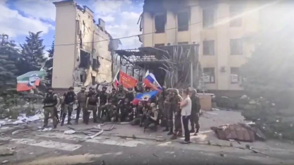 This photo taken from video provided by Ramzan Kadyrov's Official Telegram channel released on Saturday, July 2, 2022 shows Russian troops including soldiers of Chechen regiment waving Russian and Chechen republic national flags as they pose for a photo in front of a destroyed building in Lysychansk, Ukraine. (Ramzan Kadyrov's Official Telegram channel via AP)