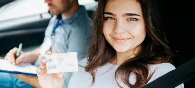 2D815X2 Attractive young woman showing driver's licence, looking at camera and smiling. Brunette woman sitting on drivers seat with fastened seat belt. Blurre