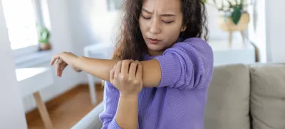 unhappy asian woman with elbow pain indoors. office syndrome health care concept. upset frowning asian lady confused looking at arms hurting sitting on couch at home in cozy bright apartment. / Foto: Stefanamer