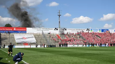 FILED - 11 June 2023, Bavaria, Unterhaching: Unterhaching coach Sandro Wagner (L) sits on the edge of the pitch while thick smoke rises behind the north stand during the the Regionalliga promotion round to the 3rd division soccer match between SpVgg Unterhaching and Energie Cottbus at Sportpark Unterhaching. The Bild paper said the council of the municipality of Unterhaching near Munich has given the green light to sell its 15,000-seat Sportpark Unterhaching stadium to Bayern for ·7.25 million (.5 million). Photo: Angelika Warmuth/dpa