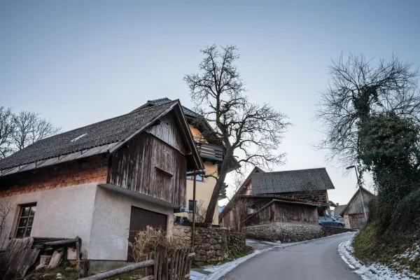 In the heart of Stara Fuzina, wooden and masonry chalets cluster along a winding lane. Textured facades, steep gabled roofs, and small windows reveal the practical alpine architecture developed for cold seasons and mountain life. Weathered boards and stone retaining walls add a sense of age and continuity, while bare trees and leftover snow signal winter. The composition highlights everyday rural character rather than tourist icons, making it ideal for illustrating Slovenian village life, vernacular building styles, heritage preservation, and the human scale of communities near Lake Bohinj. / Foto: Balkanscat Getty Images