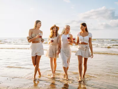 Happy women with glasses of wine walking on the beach. Celebrating bachelorette party, drinking wine at sunset. Holidays, nature, relax concept.