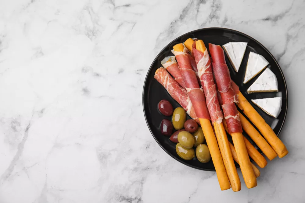 Plate of delicious grissini sticks with prosciutto, cheese and olives on white marble table, top view. Space for text / Foto: Liudmila Chernetska