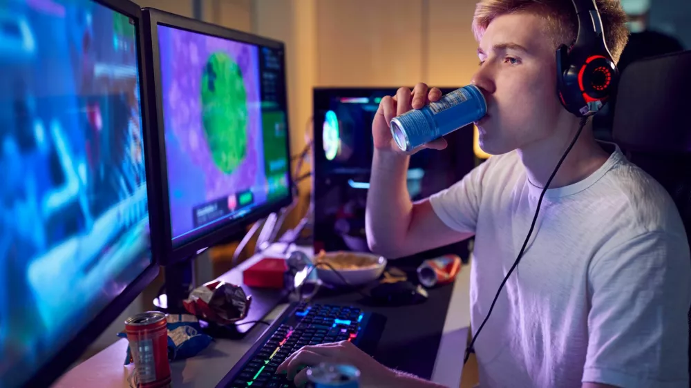Teenage Boy Drinking Caffeine Energy Drink Gaming At Home Using Dual Computer Screens At Night