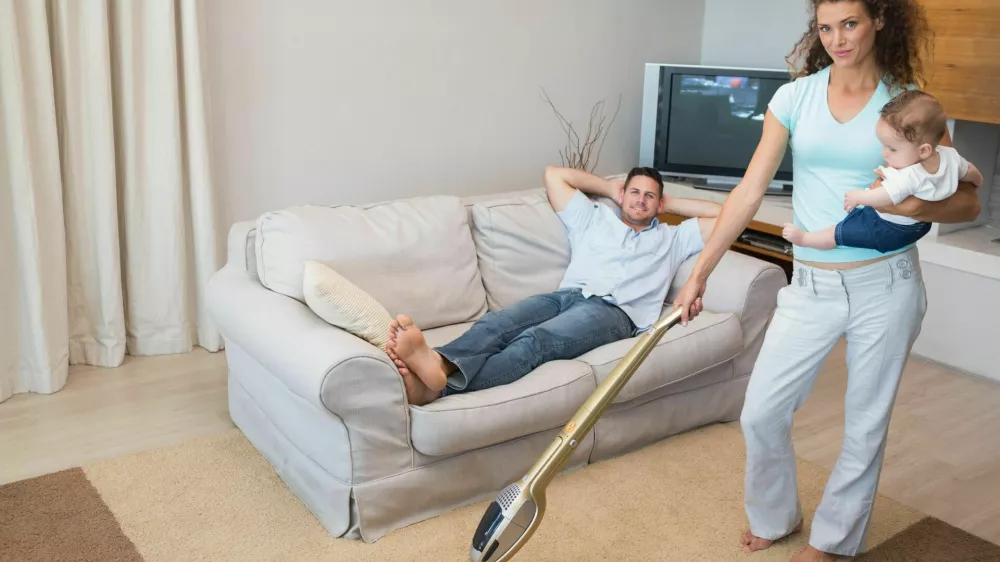 ženska čisti, či&scaron;čenje, pospravljanje, gospodinjska dela Full length portrait of woman cleaning house while carrying baby with man relaxing on sofa,Image: 184546642, License: Royalty-free, Restrictions:, Model Release: yes