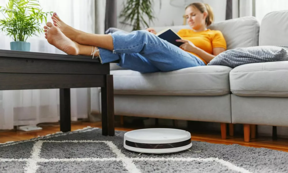 Robotic vacuum cleaner cleaning a room while a woman relaxing, reading book on the sofa. Innovative technologies for cleaning