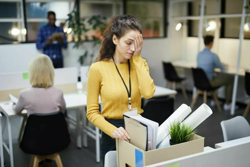 Frustrated young woman in yellow sweater standing at table and touching face with hand while packing stuff in office after dismissal / Foto: Seventyfour