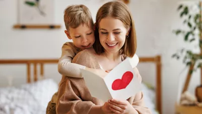 Cute little boy son congratulating his mom happy woman with Mothers day, giving her handmade greeting postcard with red heart while sitting together on bed at home. Family holidays concept / Foto: Evgenyatamanenko