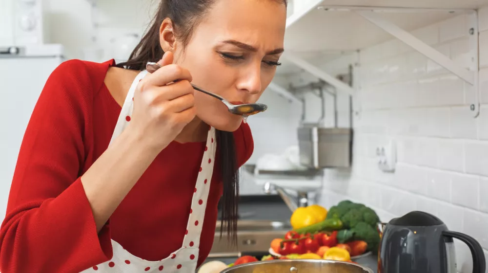 Young woman wife indoors cooking romantic dinner for husband blowing on spoon with pasta chilling out taste concentrated