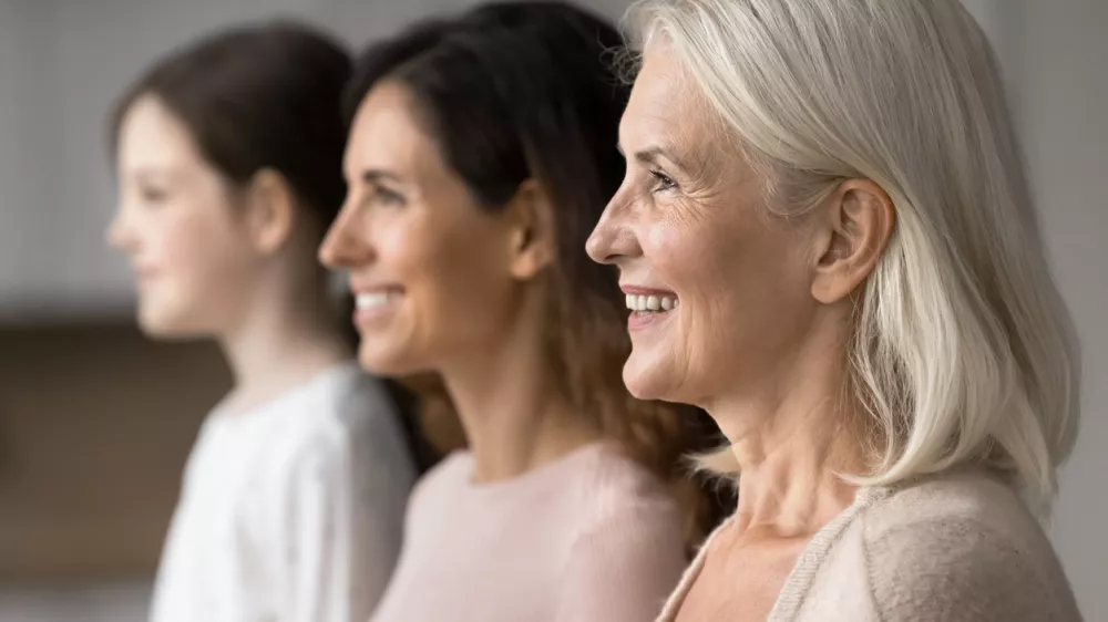 Happy attractive old 65s woman standing in row with young adult 30s daughter and little 7s granddaughter, smiling, look straight, side profile faces view. Three generations family portraits, close up / Foto: Fizkes Getty Images