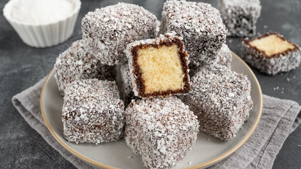 Traditional Australian Lamington cake in chocolate glaze and coconut flakes on a plate on a gray concrete background with a cup of tea. Selective focus / Foto: Irina Taskova