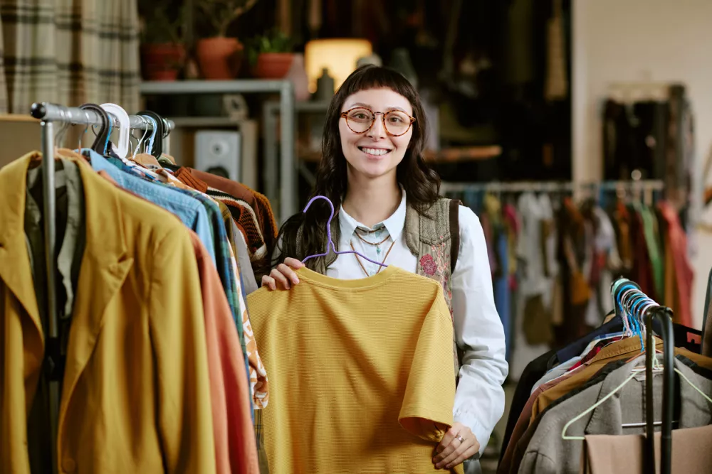 Portrait of young adult biracial woman smiling while holding second hand shirt on hanger in thrift store, surrounded by racks of used clothing, engaging in sustainable fashion shopping / Foto: Annastills Getty Images