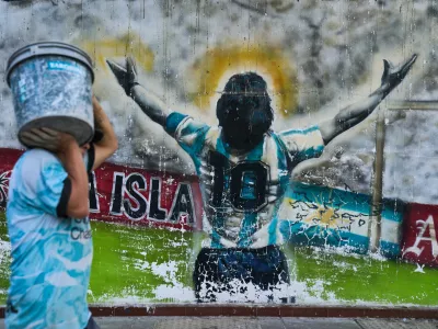 A pedestrian carries a bucket past graffiti depicting late soccer legend Diego Maradona in Buenos Aires, Argentina, Monday, April 13, 2026. (AP Photo/Rodrigo Abd)
