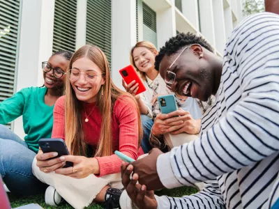 Group of smiling young multiethnic high school students using cellphones. Guys and females watching social media with their smartphones, while enjoying together sitting at the university campus lawn. High quality photo
