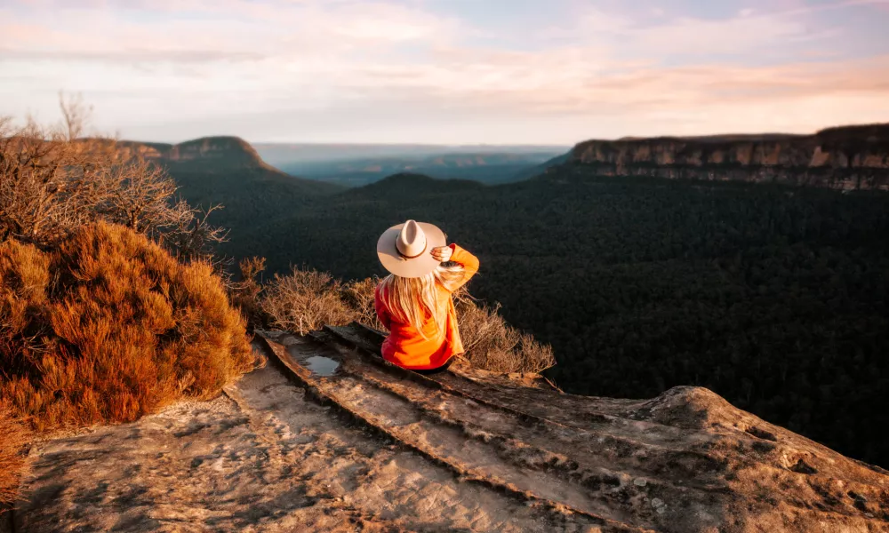 Woman sits on the edge of a cliff and looks out over the mountains in teh late afternoon