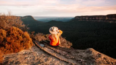 Woman sits on the edge of a cliff and looks out over the mountains in teh late afternoon