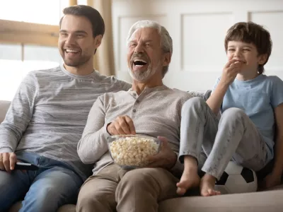 Overjoyed three generations of Caucasian men sit relax on sofa at home laugh watching video eating popcorn together. Smiling boy child with young dad and grandfather rest with snacks enjoy TV. / Foto: Fizkes