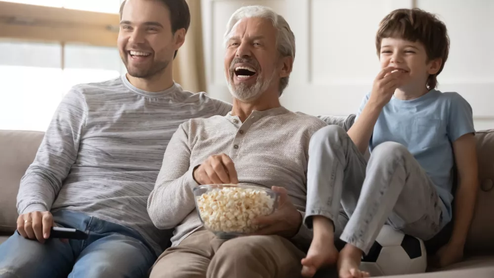 Overjoyed three generations of Caucasian men sit relax on sofa at home laugh watching video eating popcorn together. Smiling boy child with young dad and grandfather rest with snacks enjoy TV. / Foto: Fizkes