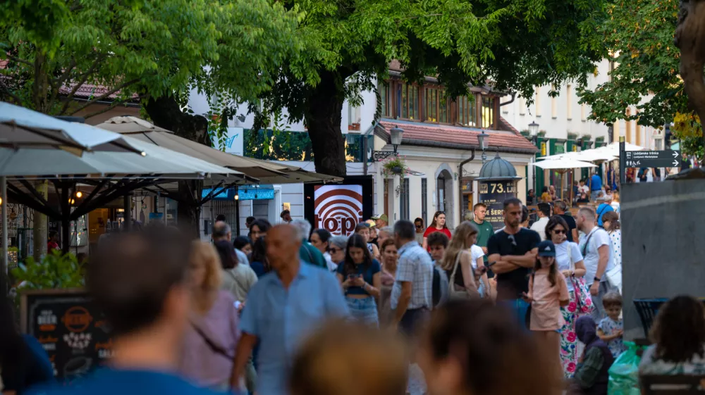 Capturing the lively atmosphere of a European summer, this shot shows a bustling pedestrian street in Ljubljana's historic center. Crowds of tourists and locals stroll past outdoor cafes and shops shaded by large trees. The warm natural light and vibrant urban energy make it ideal for travel guides, city lifestyle features, and tourism marketing showcasing popular destinations in Slovenia. / Foto: Aleksei Gorovoi Getty Images
