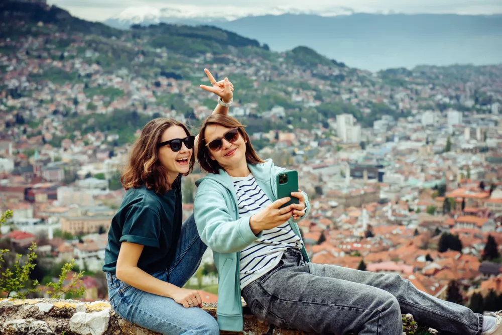 Two cheerful traveler girls in dark glasses are having fun and taking pictures against the background of Sarajevo, a beautiful European city in the mountains.
