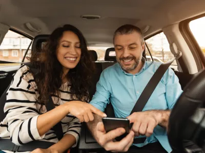 Short Road Route. Portrait of smiling couple sitting inside luxury car pointing fingers at smart phone, cheerful male driver holding cell, showing choosing location, using digital map application