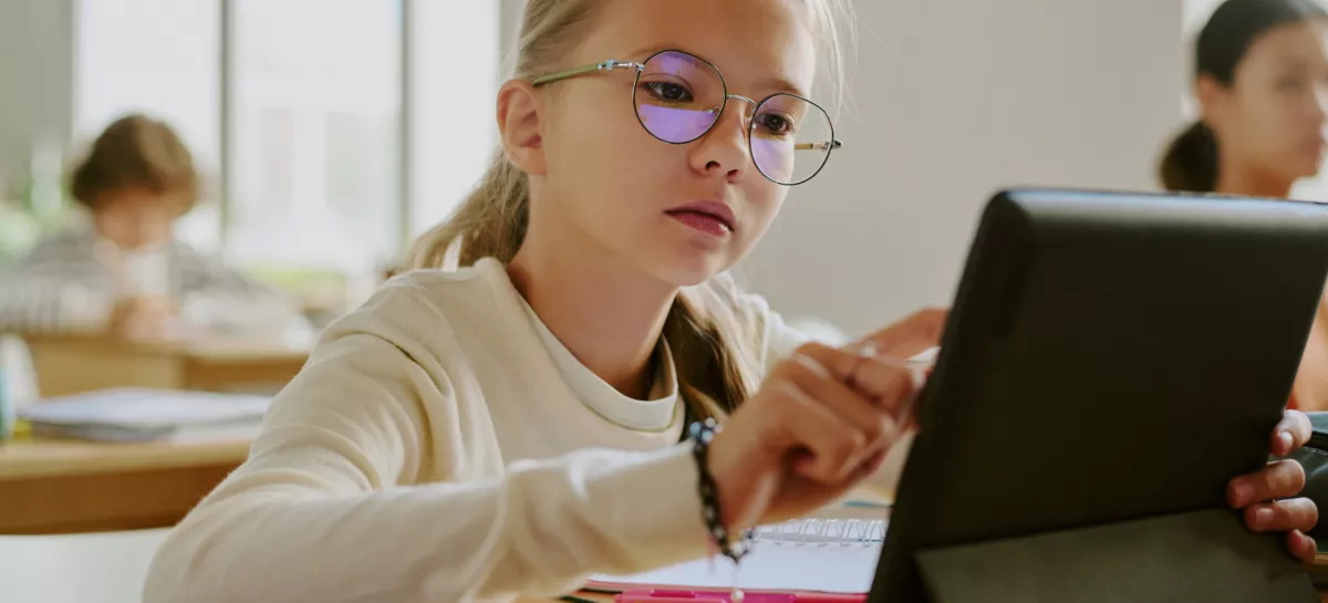 Young girl wearing glasses sitting at wooden desk using tablet in classroom surrounded by fellow students in bright learning environment / Foto: Annastills Getty Images