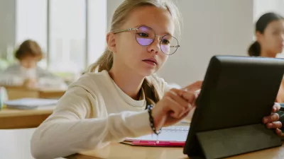 Young girl wearing glasses sitting at wooden desk using tablet in classroom surrounded by fellow students in bright learning environment / Foto: Annastills Getty Images