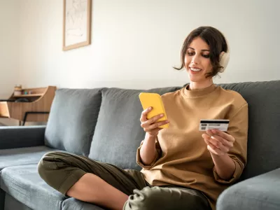 Young freelance woman using cellphone smartphone paying app and credit debit card for online shopping sitting at home on sofa. Latin girl freelancer adult female student doing money transfer by phone