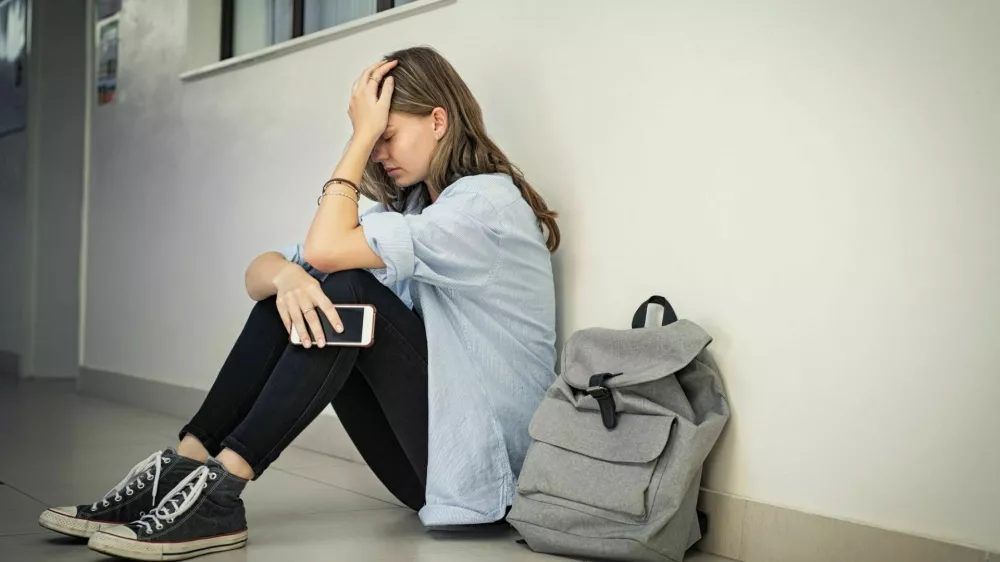 Upset and depressed girl holding smartphone sitting on college campus floor holding head. University sad student suffering from depression sitting on floor at high school. Lonely bullied teen in difficulty with copy space.