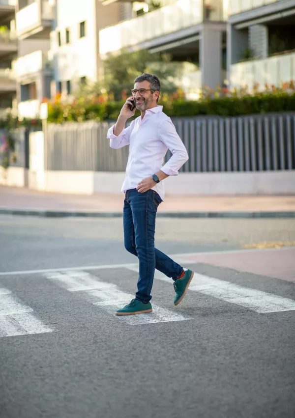 Middle-aged man in a white pink shirt and jeans, smiling while talking on his phone and crossing a city street at a pedestrian crosswalk, with modern buildings in the background / Foto: Todayistuesday