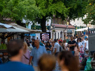 Capturing the lively atmosphere of a European summer, this shot shows a bustling pedestrian street in Ljubljana's historic center. Crowds of tourists and locals stroll past outdoor cafes and shops shaded by large trees. The warm natural light and vibrant urban energy make it ideal for travel guides, city lifestyle features, and tourism marketing showcasing popular destinations in Slovenia.