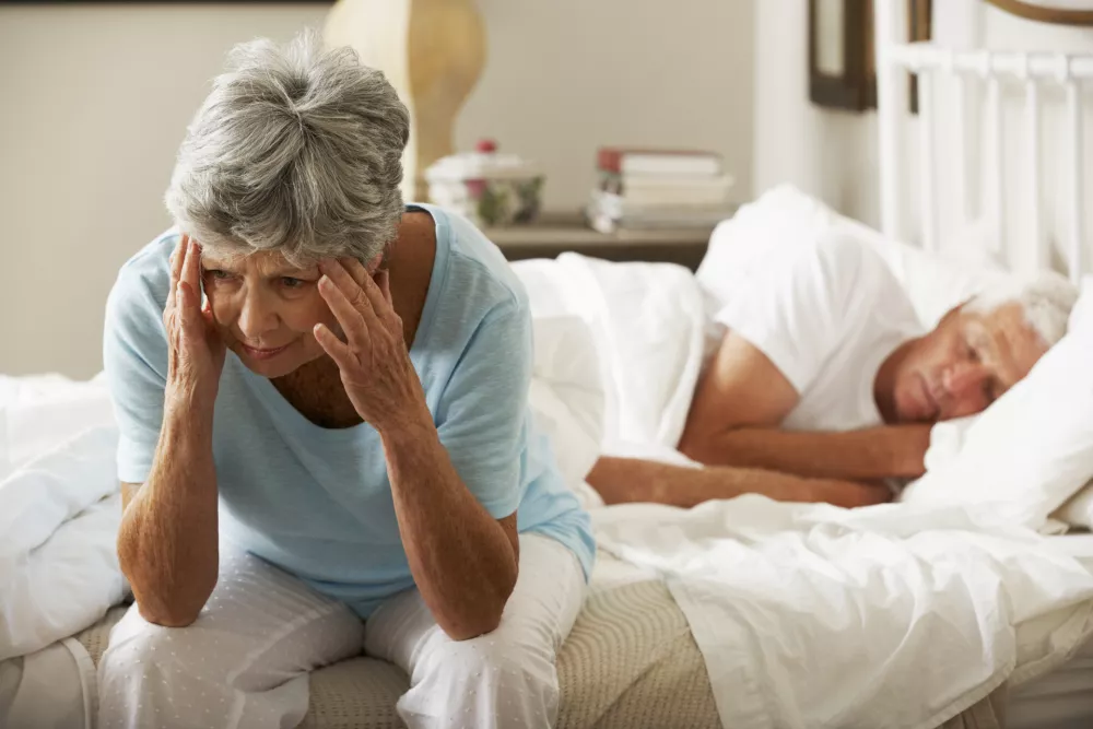 Worried Senior Woman Sits On Bed Whilst Husband Sleeps. / Foto: Monkeybusinessimages