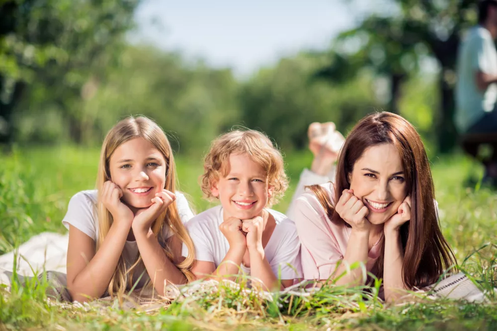 Mother and two children lie on a picnic blanket in a green park, smiling and enjoying a sunny summer day together. Warm family bonding, playful mood and relaxed outdoor lifestyle. / Foto: Deagreez