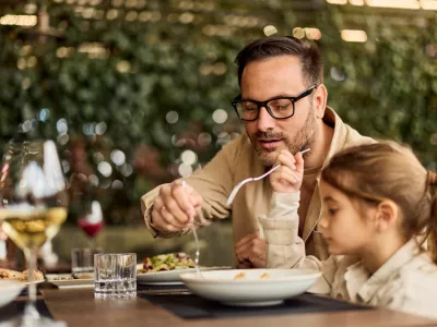Father and young daughter share a relaxed lunch at an outdoor restaurant, passing food and enjoying quality family time in a casual, warm setting ideal for lifestyle, parenting, and hospitality stories. / Foto: Nortonrsx Getty Images