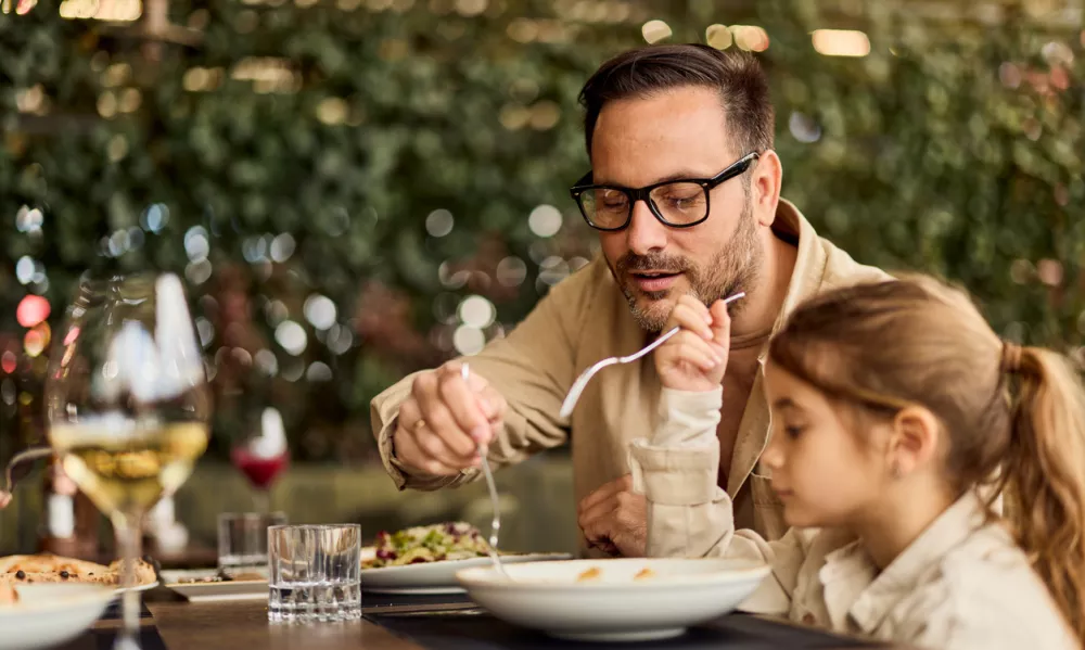 Father and young daughter share a relaxed lunch at an outdoor restaurant, passing food and enjoying quality family time in a casual, warm setting ideal for lifestyle, parenting, and hospitality stories. / Foto: Nortonrsx Getty Images