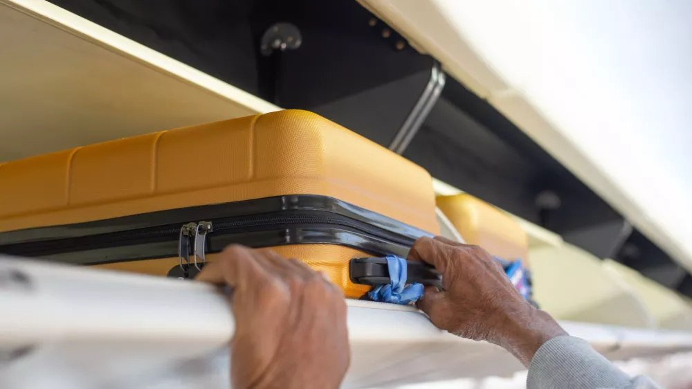 A person's hands are seen placing or retrieving a yellow hard-shell suitcase from an airplane's overhead storage compartment.