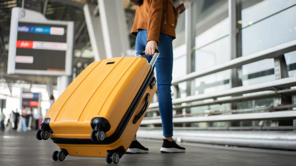 Close up of unrecognized woman tourist walking with her luggage through international airport terminal during holiday trip, female tourist rolling suitcase alone.