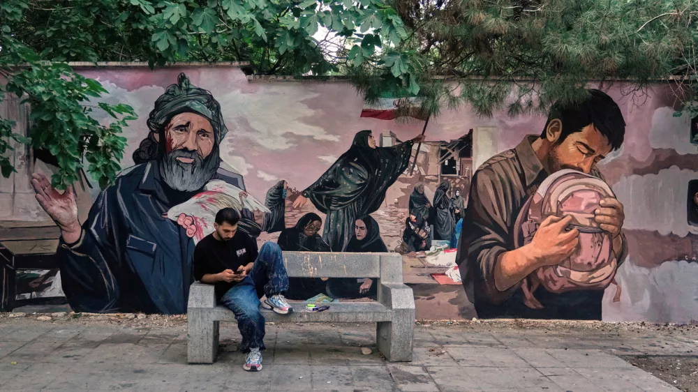 A man works on his smart phone in front of a mural honoring schoolchildren killed in a Feb. 28 strike in the southern Iranian city of Minab that was likely caused by U.S. airstrikes, in northern Tehran, Monday, June 15, 2026. (AP Photo/Vahid Salemi)