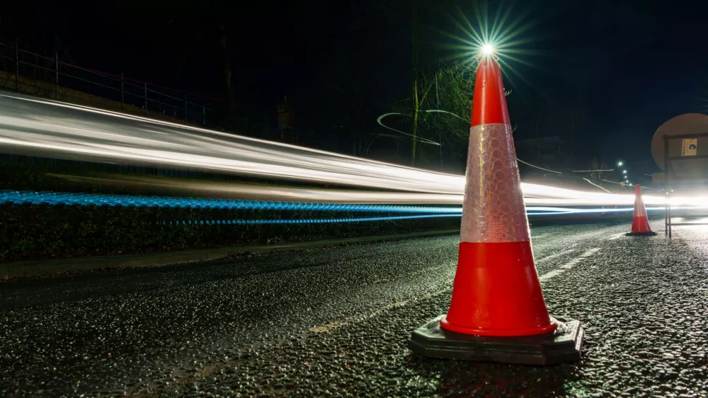 Traffic cone on a road at night with light trails from passing vehicles.