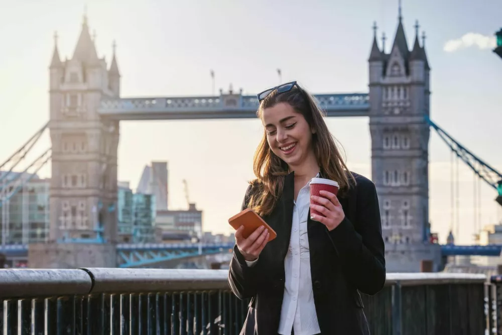 A young business woman walking with a cup of coffee and looking at her mobile phone in front of the City skyline of London / Foto: Shansche