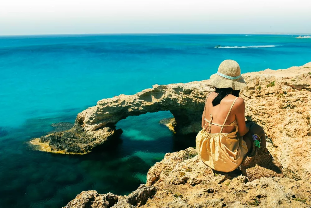 Woman tourist sit on viewpoint sightseeing Cyprus greek island visit stone arch or bridge of Love. Famous holiday destination in mediterranean seacoast / Foto: Eval