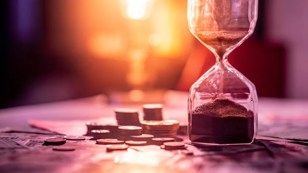 Sand running through the shape of hourglass on table with banknotes and coins of international currency. Time investment and retirement saving. Urgency countdown timer for business deadline concept / Foto: Zephyr18 Getty Images/istockphoto