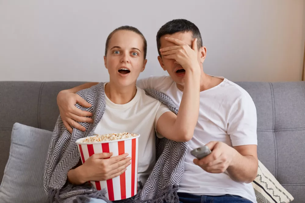Portrait of impressed woman watching horror film with man, shocked wife covered husband's eyes with palm, forbidden content on tv, couple sitting on sofa wrapped in blanket. / Foto: Sementsovalesia