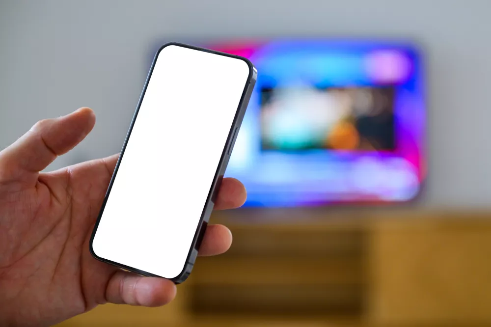Smartphone mockup screen, man holding mobile phone in front of television set, tv remote app and tv program listing concept, selective focus / Foto: Bitsandsplits